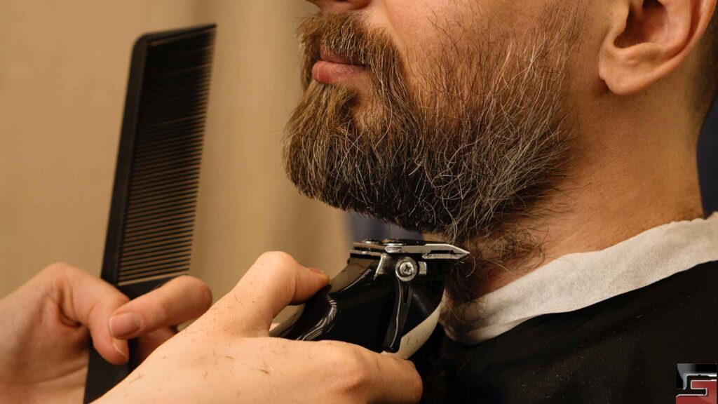 A man gets his thick, brown beard trimmed by a barber.